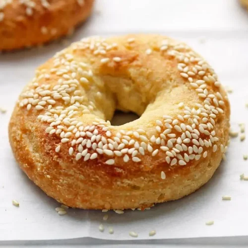 A stack of golden brown Keto Bagels with sesame seeds, with one bagel sliced and spread with cream cheese in the foreground.