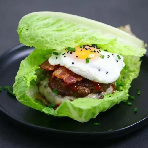 A keto breakfast burger stacked in a crisp lettuce wrap with a juicy beef patty, melted swiss cheese, bacon, and a fried egg.