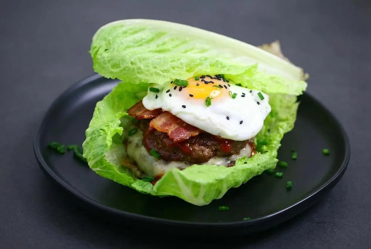 A keto breakfast burger stacked in a crisp lettuce wrap with a juicy beef patty, melted swiss cheese, bacon, and a fried egg.