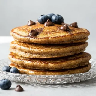 A stack of fluffy keto peanut pancakes on a light grey plate, topped with a melting pat of butter, fresh blueberries, and raspberries.