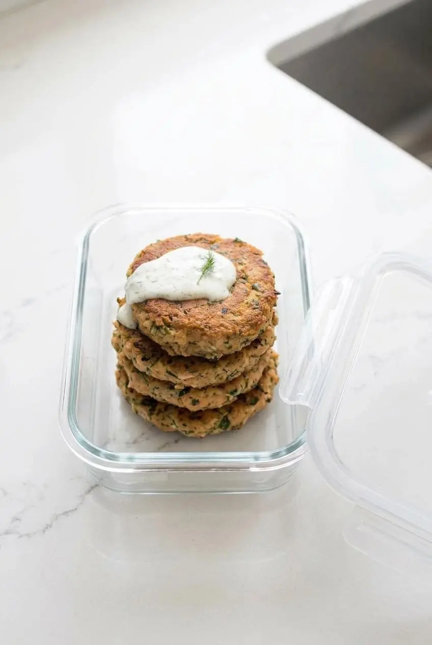 Four cooked keto salmon patties stacked inside a glass airtight container for meal prep storage.
